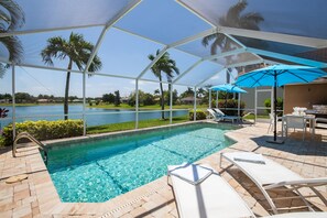 Piscina con vista a un lago, rodeada de palmeras y un patio cubierto con sillones reclinables y sombrillas.
