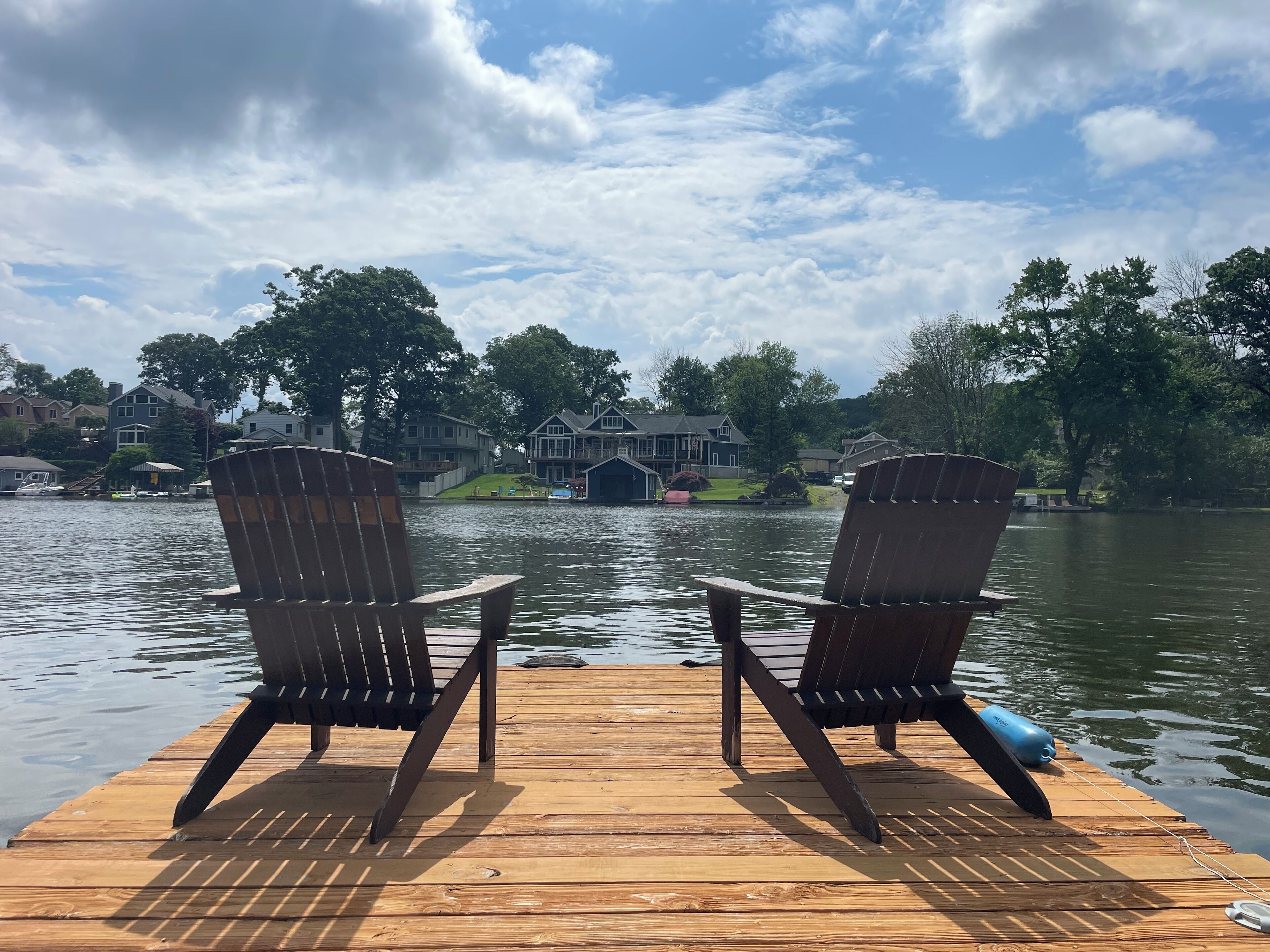 Two wooden Adirondack chairs on a dock with a lake and houses in the background.