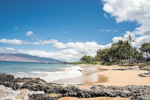 Uma praia com pedras em primeiro plano, céu claro e montanhas ao longe.