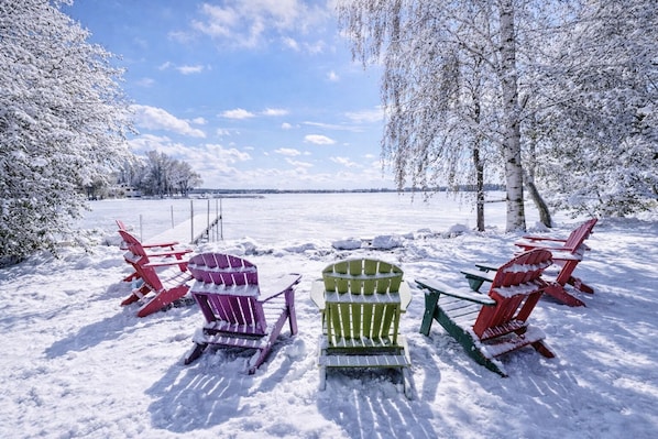 A snow-covered outdoor seating area with Adirondack chairs and a table, surrounded by trees and a view of a frozen lake.