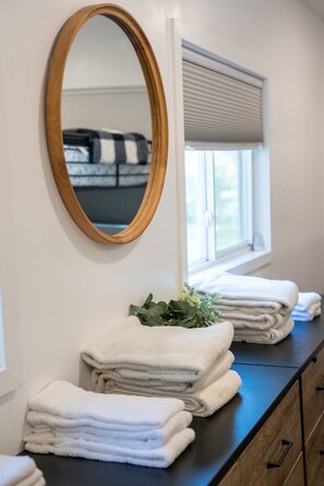 A bathroom with a wooden-framed mirror, a window with blinds, and a stack of white towels on a dark countertop.