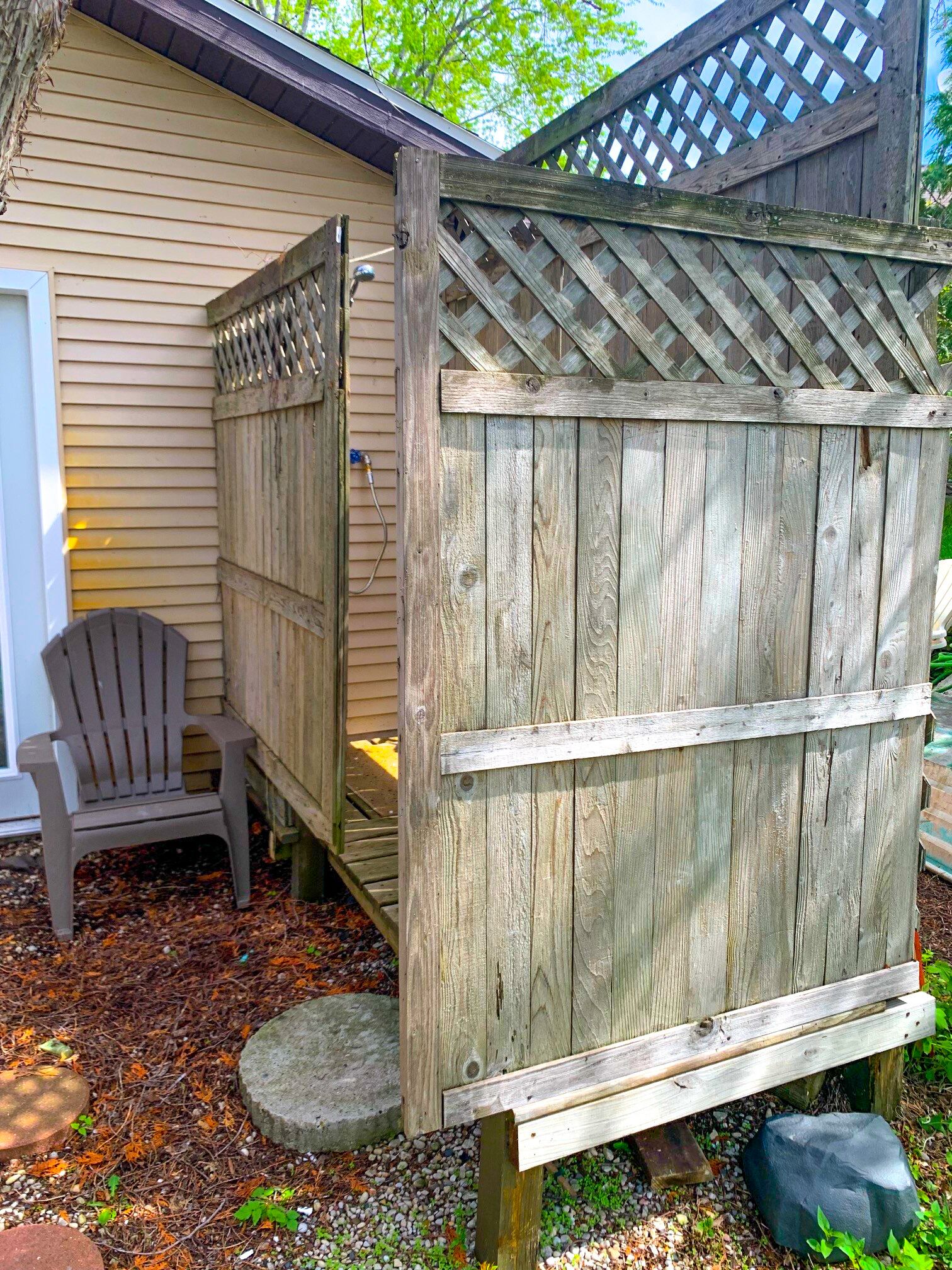 Una valla de madera con parte superior de celosía, una silla Adirondack de plástico y un pequeño banco de piedra.