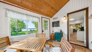 Une pièce aux murs en bois, dotée d’une grande fenêtre donnant sur une pelouse et un lac. On y trouve une table en bois avec des chaises, un meuble turquoise et un panneau d’accueil.