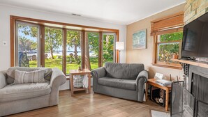 Un salon avec deux canapés, une table basse et un téléviseur. De grandes fenêtres offrent une vue sur un jardin et un plan d’eau.