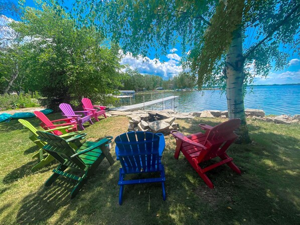 An outdoor seating area with Adirondack chairs arranged around a fire pit, overlooking a body of water.