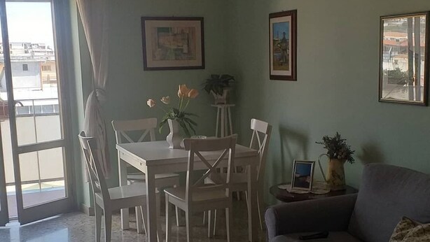 Sala de estar con una mesa de comedor, sillas, un sofá y una mesa de centro. Hay dos cuadros en la pared, una planta en maceta y una ventana con cortinas.