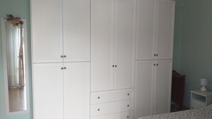 Una habitación con un armario blanco grande, una mesita de noche pequeña con lámpara y una ventana con cortinas.