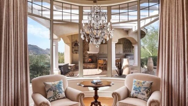 A room with two armchairs, a table, a chandelier, and large windows offering a view of a fireplace and outdoor area.