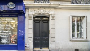 A building facade with a black double door, a window with decorative iron bars, and a blue storefront with signage and posters.
