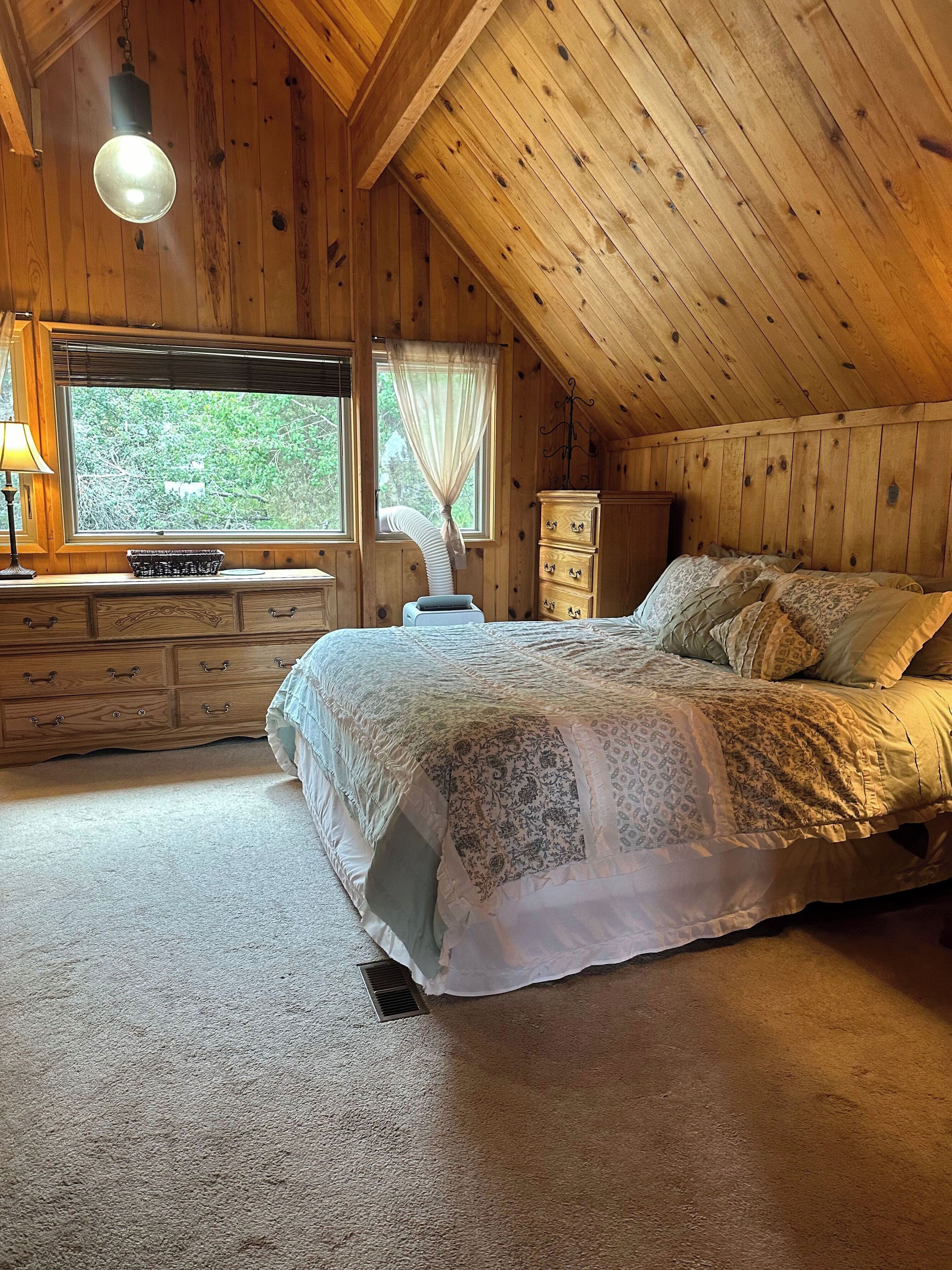 A cozy bedroom with a wooden ceiling, a large bed with patterned bedding, a dresser, and a window with curtains.
