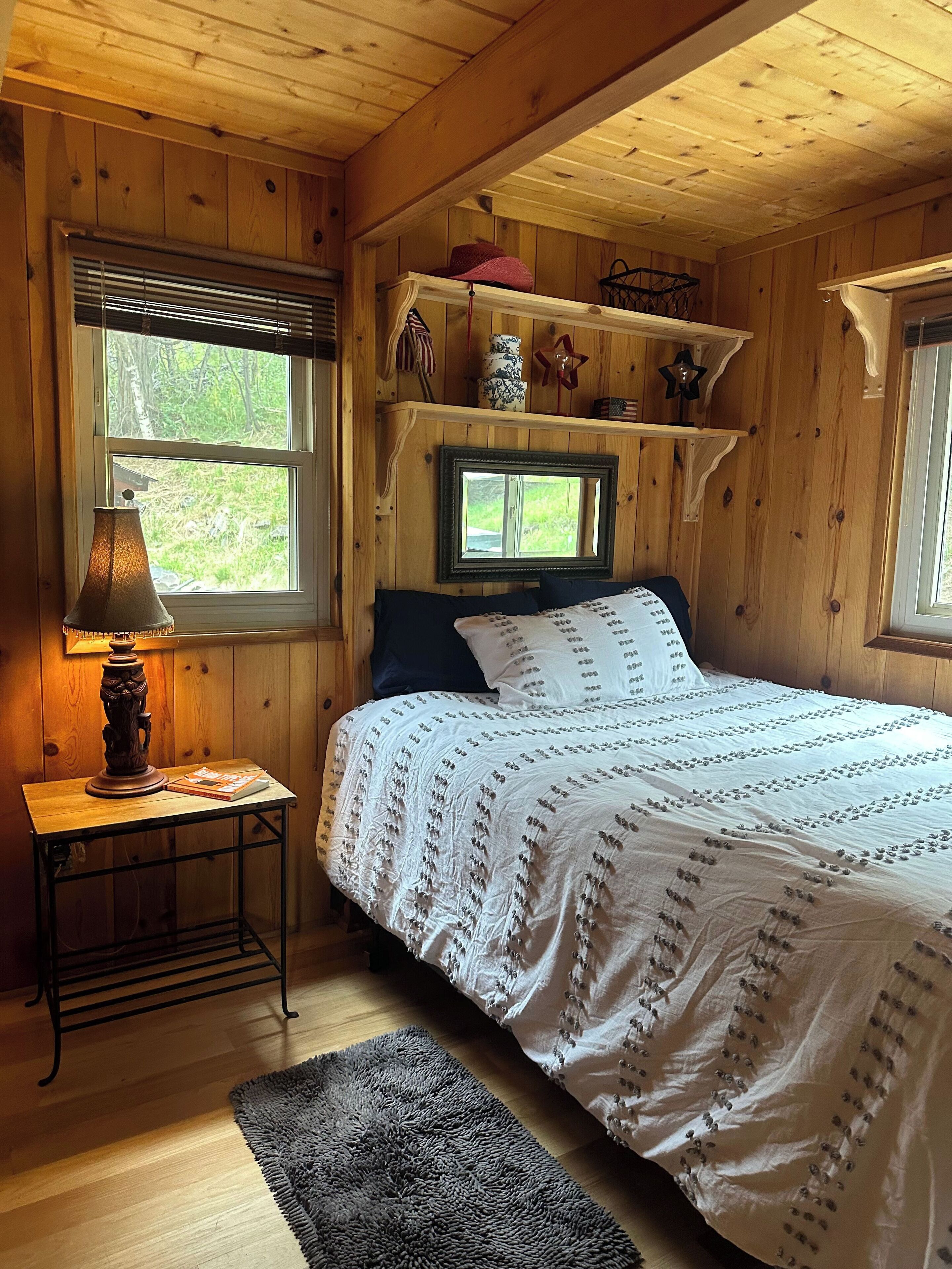 Un dormitorio de cabaña de madera con una cama, una mesita de noche con una lámpara y estanterías sobre la cama.