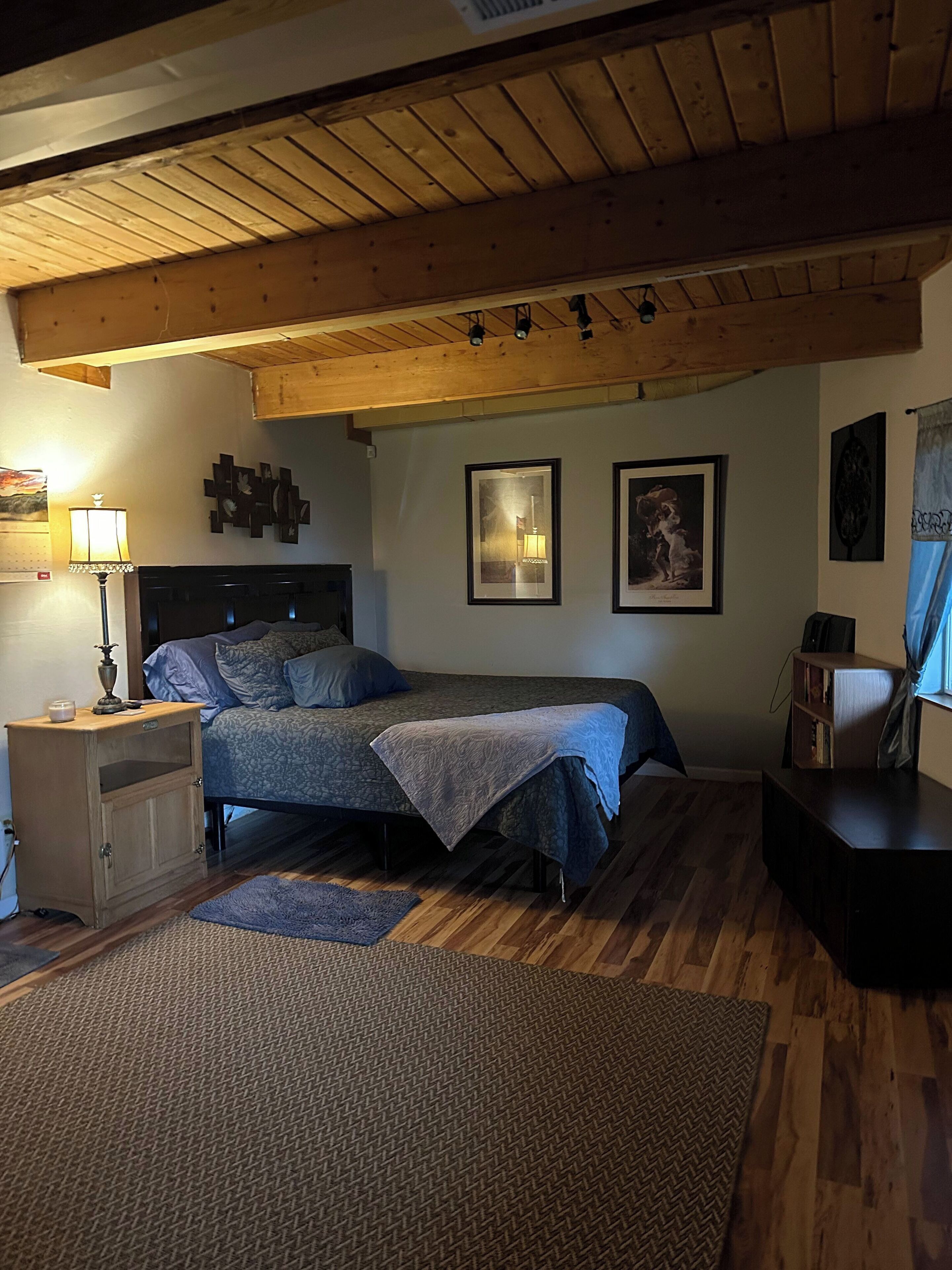 Ein Schlafzimmer mit Holzboden, einem Bett mit gemustertem Überwurf, einem Nachttisch mit Lampe und gerahmten Bildern an der Wand.