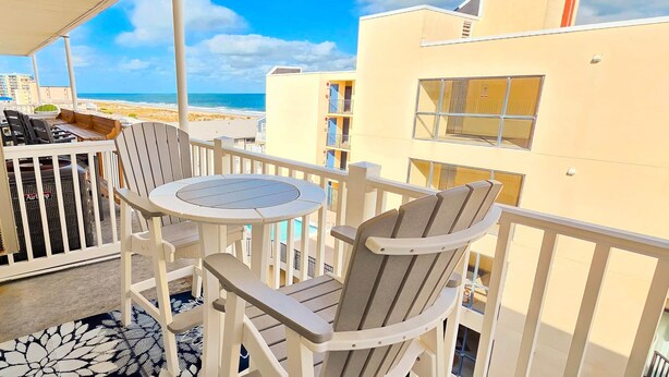 A balcony with a table and chairs, overlooking a beach and a pool.
