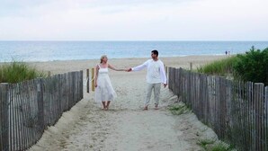 A couple holding hands on a sandy path with a wooden fence on either side, leading to a beach with the ocean in the background.