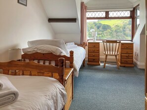 A bedroom with a bed, a desk, a chair, and a window with a view of greenery.