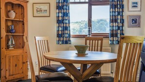 Een eethoek met een ronde houten tafel, twee houten stoelen met zwarte kussens, een houten kast met decoratieve spullen, en een raam met blauwe gordijnen.