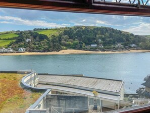 A view of a waterfront property with a wooden deck, a glass balustrade, and a backdrop of green hills and houses.