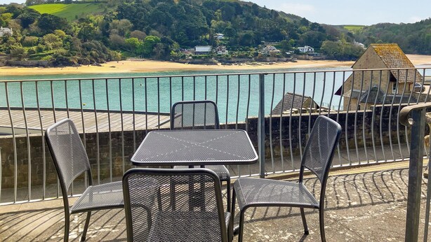Een terras met metalen stoelen en een tafel, met uitzicht op een strand en heuvels.