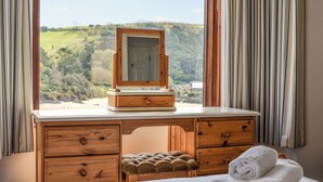 Een houten wastafel met spiegel, een bed met wit beddengoed en uitzicht op een groen landschap door het raam.