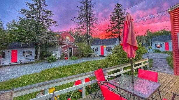 A wooden deck with red chairs and a table, overlooking a row of cabins with red doors and windows. - Open Hearth Inn Suite 2 - 10 min to Acadia! (Trenton)