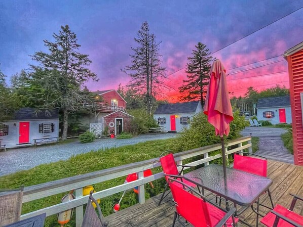A wooden deck with red chairs and a table, overlooking a row of cabins with red doors and windows.