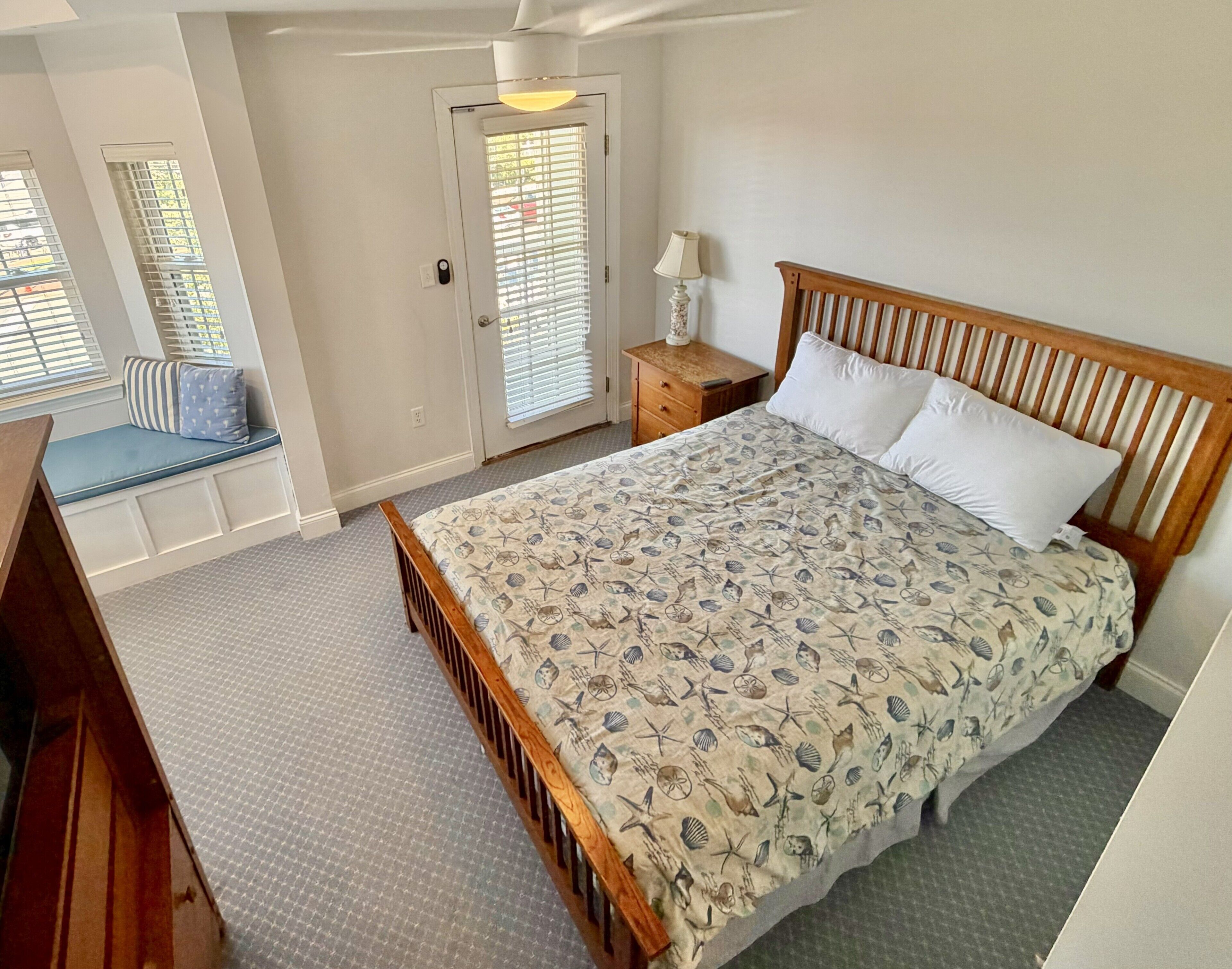 Une chambre à coucher comprenant un lit, une table de chevet, une lampe, un banc avec des coussins et une porte avec des stores.