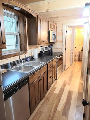 A compact kitchen with wooden cabinets, a double sink, a microwave, and a dishwasher. - Queen of Hearts. Secluded Hocking Hills cabin. (Rockbridge)