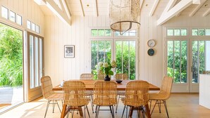 A dining area with a wooden table and wicker chairs, large windows, and a hanging light fixture.