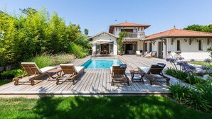 A poolside area with wooden deck chairs, a pool, and a two-story house with a balcony.