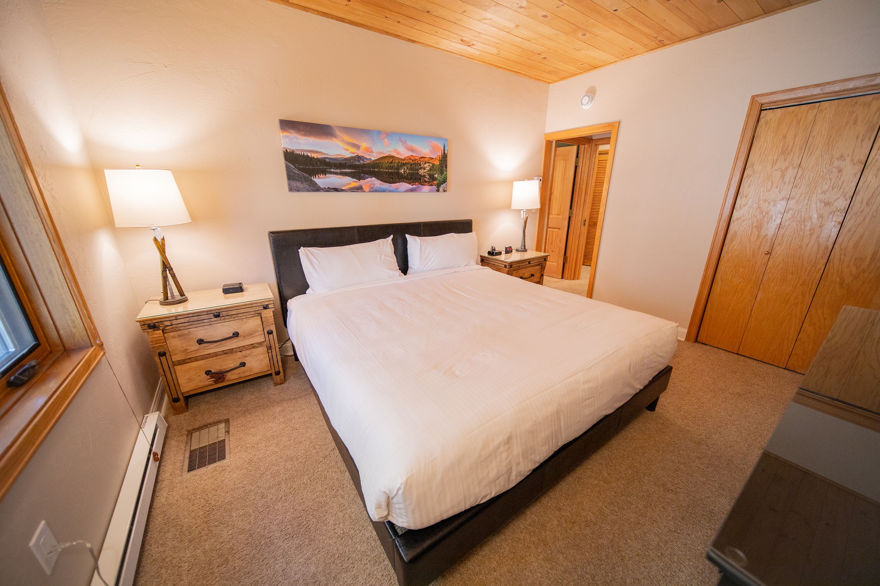 Een slaapkamer met een groot bed, een houten hoofdbord, twee nachtkastjes met lampen, een bijzettafeltje, een houten garderobe en een schilderij aan de muur.