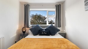 A bedroom with a bed, a window with curtains, a bedside table with a lamp, and a pillow with text. - Ty Gwyn - Hot Tub Parking Dog Friendly (Saundersfoot)