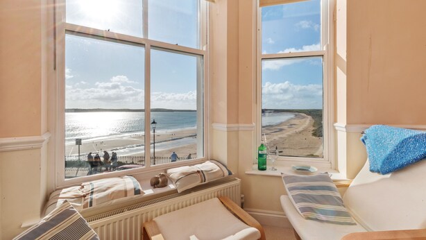 A room with a large window overlooking a beach, a bed, a small table with a bottle and glasses, and a bench with a towel.