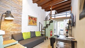 A cozy living room with a sofa, a small table, and a clear chair. There is a staircase leading to an upper level, and a fire extinguisher mounted on the wall. - Casina Magnoli (Florence)