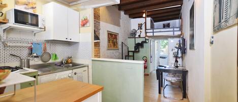 A compact kitchen with white cabinets, a microwave, and a stove. There is a small island with a sink and a stool. The kitchen has wooden flooring and a stone accent wall.