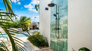 A modern outdoor shower area with a tiled wall, a pool, and lush greenery. - Massive Oceanfront Villa - Spa - Sauna - Gameroom - Malmok! (Noord)