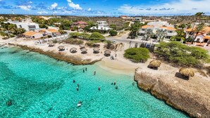 An aerial view of a coastal area with clear turquoise waters, a sandy beach, and several buildings with red-tiled roofs. - Massive Oceanfront Villa - Spa - Sauna - Gameroom - Malmok! (Noord)