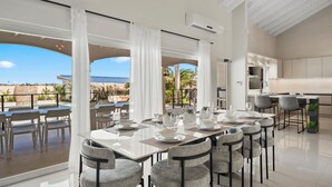 A modern dining area with a view of the beach, large windows, and a kitchen area. - Massive Oceanfront Villa - Spa - Sauna - Gameroom - Malmok! (Noord)