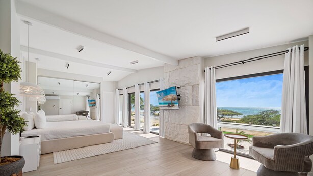 A modern hotel room with a large bed, a seating area with two chairs, a small round table, and a large window offering a view of the ocean. - Massive Oceanfront Villa - Spa - Sauna - Gameroom - Malmok! (Noord)