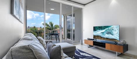Ein modernes Wohnzimmer mit einem großen Flachbildfernseher, einem grauen Sofa und einer Glastür, die zu einem Außenbereich mit Blick auf Palmen und Gebäude führt.
