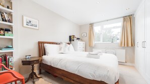 A bedroom with a wooden bed, white bedding, a bedside table, a chair, a window with curtains, a radiator, and a bookshelf. - Supreme Hampstead Home Next to Hampstead Heath by Underthedoormat (London)