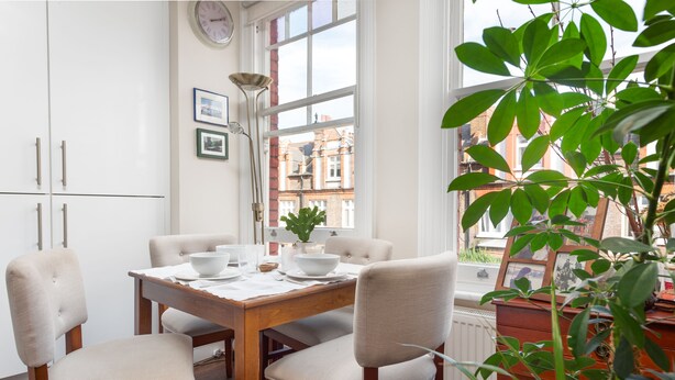 A dining area with a wooden table set for four, white chairs, a clock on the wall, and a view of buildings through a window. - Supreme Hampstead Home Next to Hampstead Heath by Underthedoormat (London)