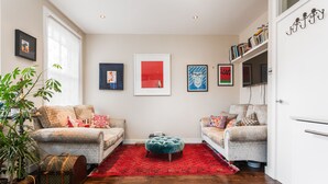 A living room with two sofas, a red rug, and a small ottoman. There are framed artworks on the wall and a potted plant on the left. - Supreme Hampstead Home Next to Hampstead Heath by Underthedoormat (London)
