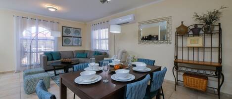 Un comedor con una mesa preparada para cuatro personas, rodeada de sillas. Se observa un sofá y una mesa de centro al fondo.