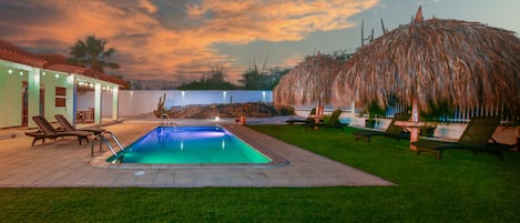 A swimming pool with lounge chairs, a thatched-roof structure, and a house with a white exterior.