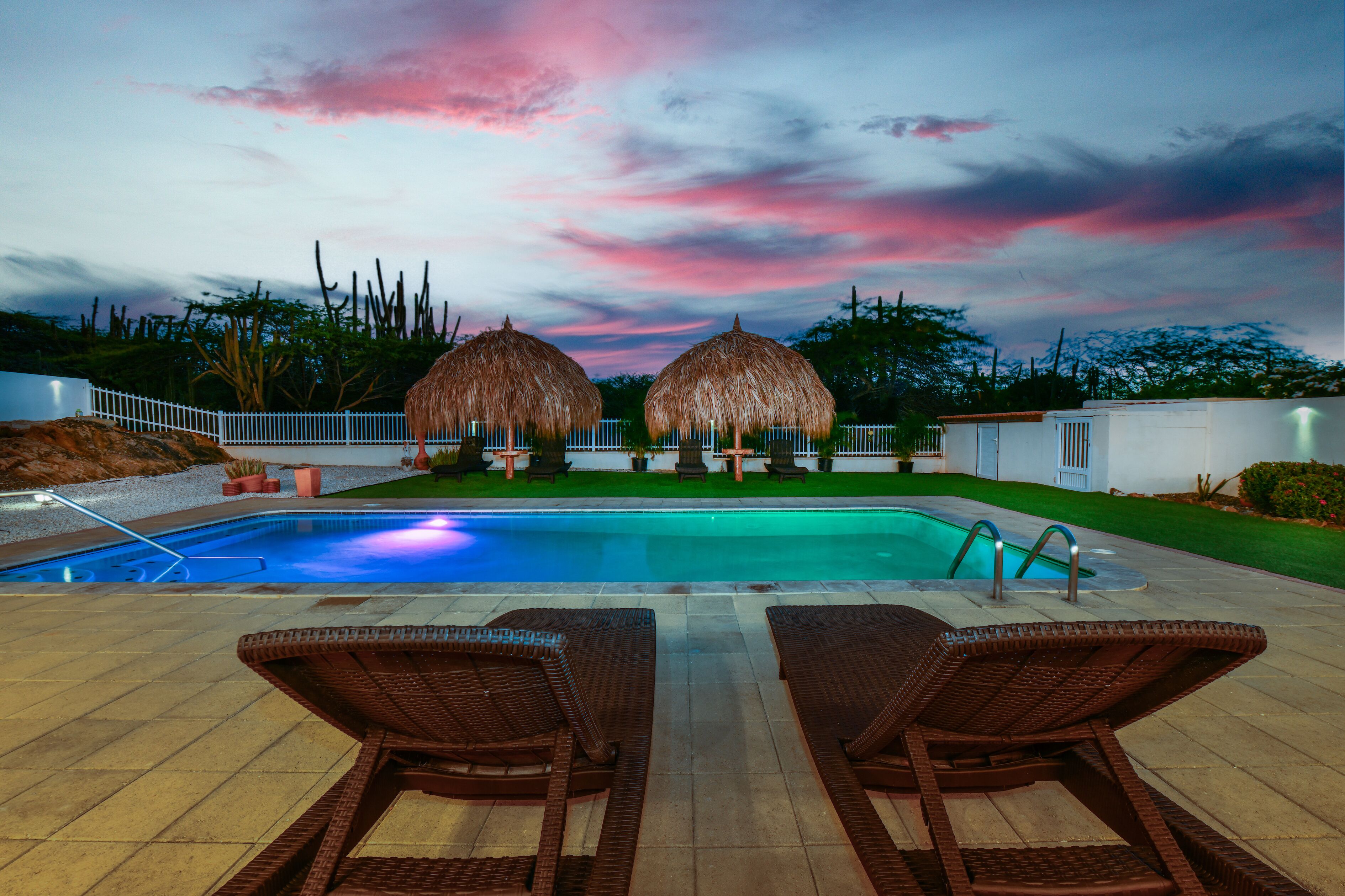 A swimming pool with lounge chairs and thatched umbrellas, surrounded by a white fence and greenery.