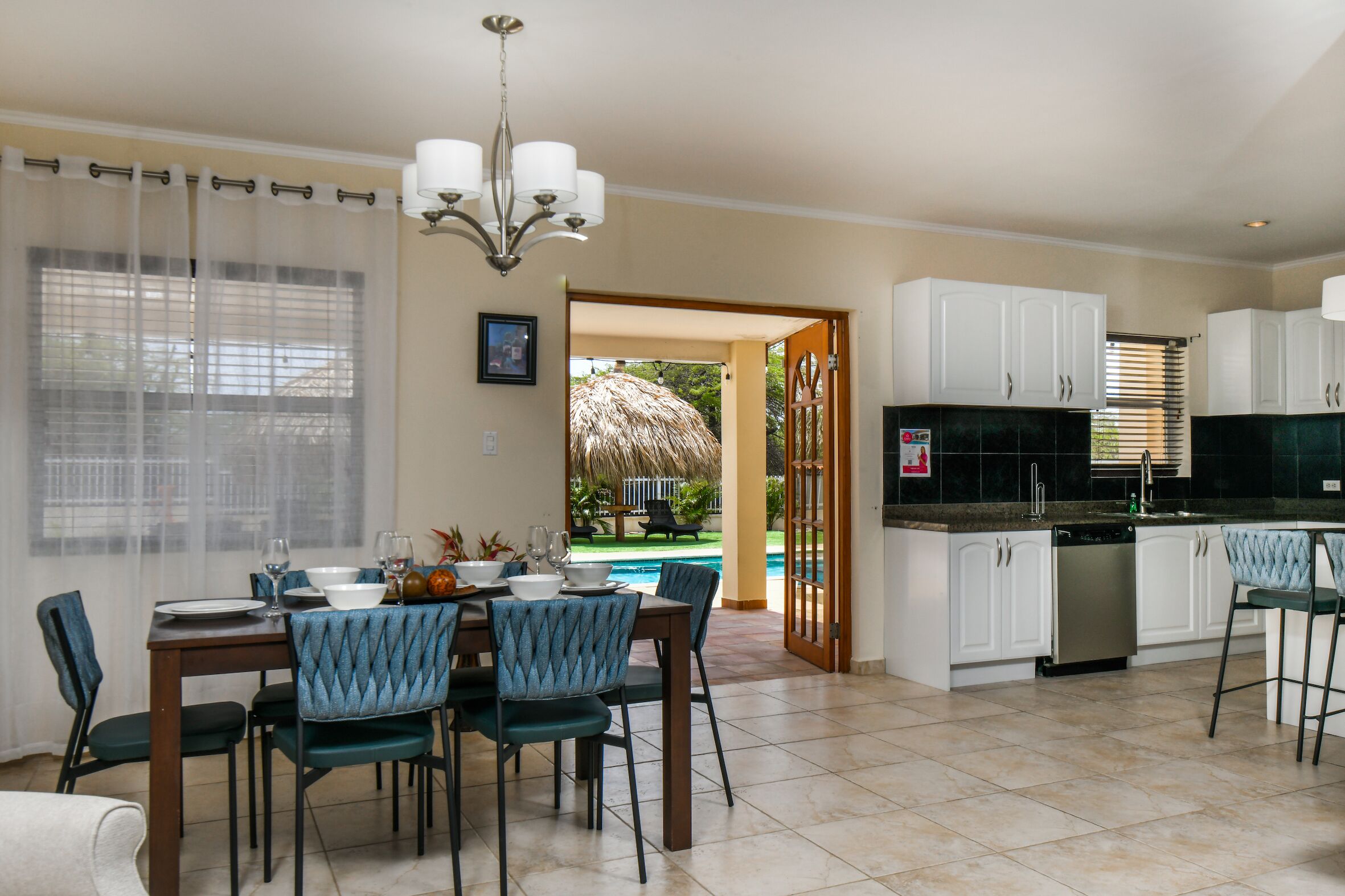 A dining area with a table set for four, a kitchen with white cabinets, and a view of a swimming pool.