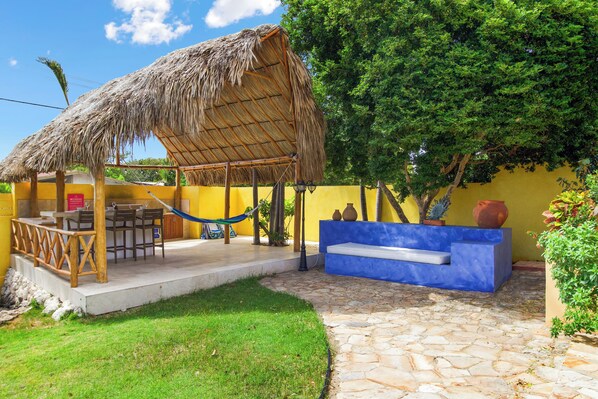 A thatched-roof outdoor seating area with a stone-tiled patio, a blue bench, and a yellow wall.