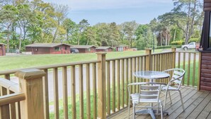 A wooden deck with a metal table and chairs, overlooking a grassy area with buildings and trees. - Lavender Lodge (Saltburn-by-the-Sea)