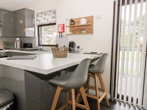 A modern kitchen with a large island, grey bar stools, a microwave, a coffee maker, and a window with blinds. - Lavender Lodge (Saltburn-by-the-Sea)