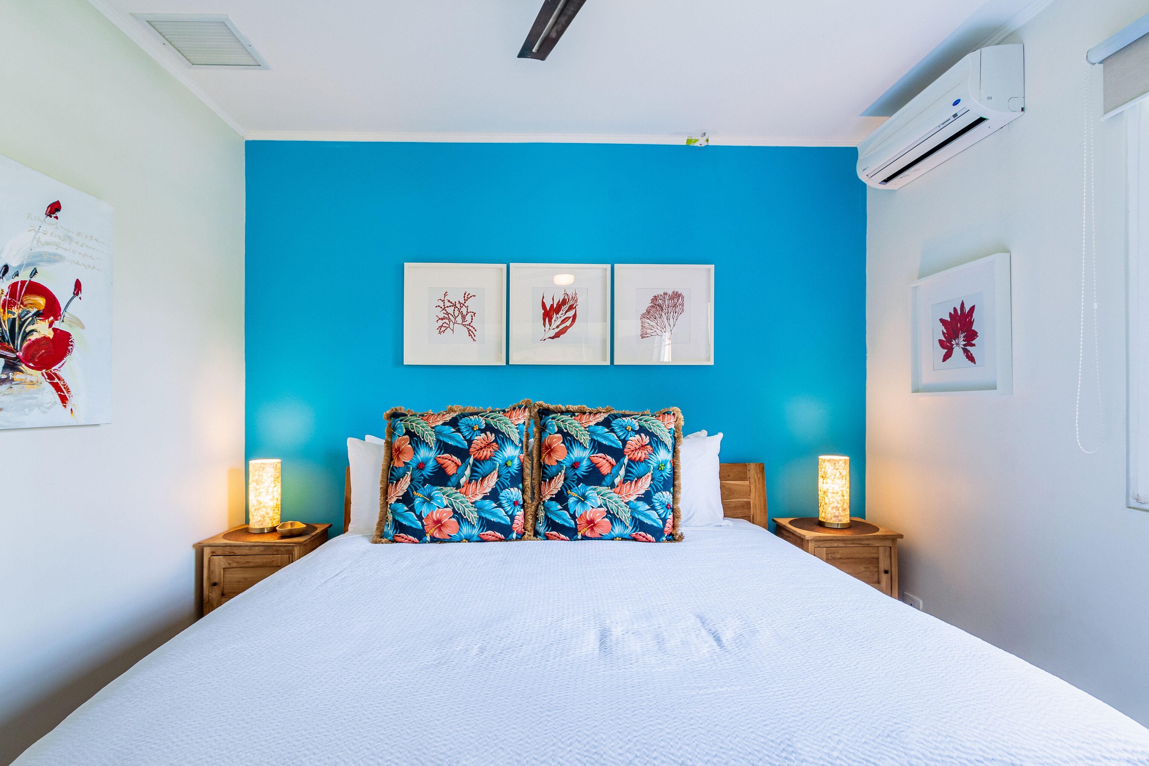 Ein Schlafzimmer mit blauer Akzentwand, einem Bett mit weißer Bettwäsche, zwei Nachttischlampen und drei gerahmten Kunstwerken über dem Bett.